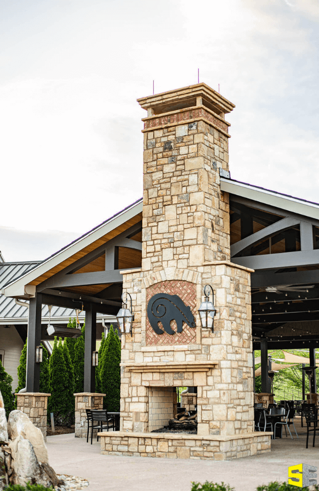 Large fireplace with a concrete image of a bear outside a constructed, wooden bodega with landscaped trees surrounding it