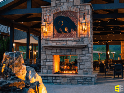 A fireplace centered under a gazebo like structure, with a bear symbol in the hearth, warm lights on either side of it.