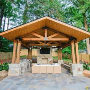 The finished product of a residential outdoor gazebo, fit with a concrete frilling area and a concrete fireplace, a large flat screen tv mounted above it