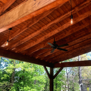 A ceiling fan mounted to the wooden structure of an outdoor gazebo ceiling, surrounded by decorative light bulb like lighting