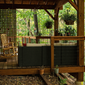 A backyard wooden gazebo area, with an outdoor couch and rocking chair, surrounded by hanging plants and the forest around it