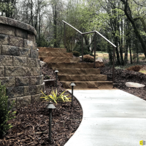 A concrete walkway, leading to stone steps that wrap around a retaining wall with natural landscaping surrounding the scene